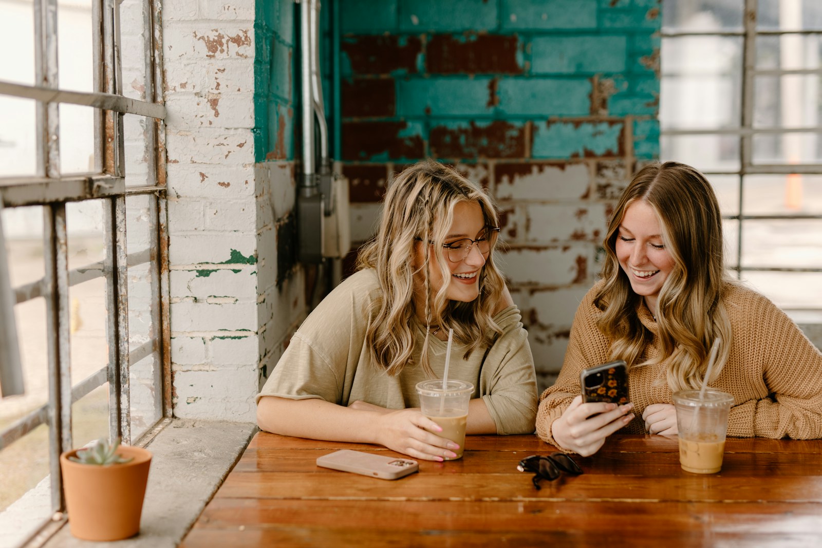 Dos mujeres sonríen mientras miran juntas un celular junto a una ventana.
